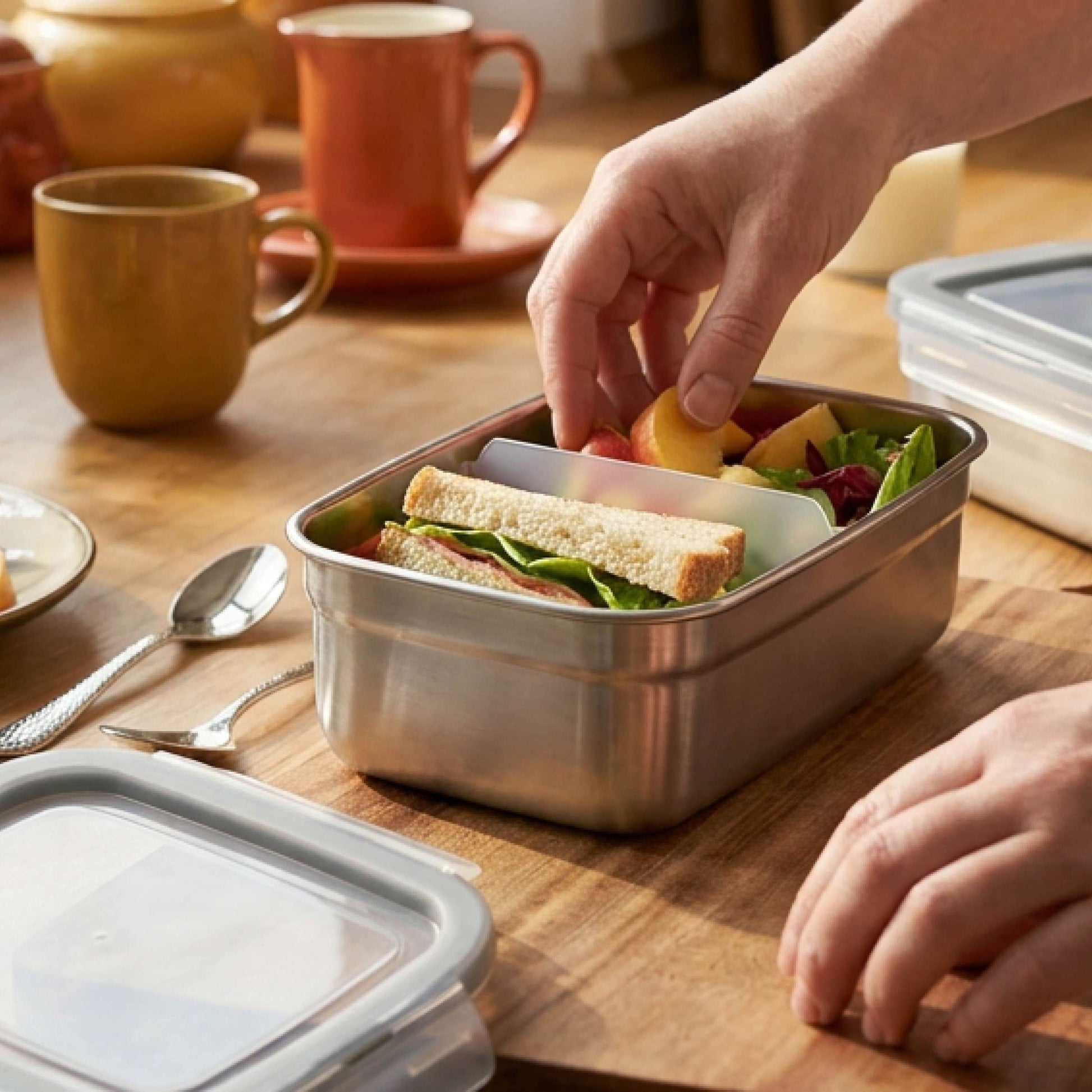 Jemand packt ein Sandwich, Obst und Salat in die „Kuppels SELECTION Frischhaltedose 2-teilig“ auf einem Holztisch, neben der Becher, Teller, Löffel und der spülmaschinenfeste Deckel liegen.