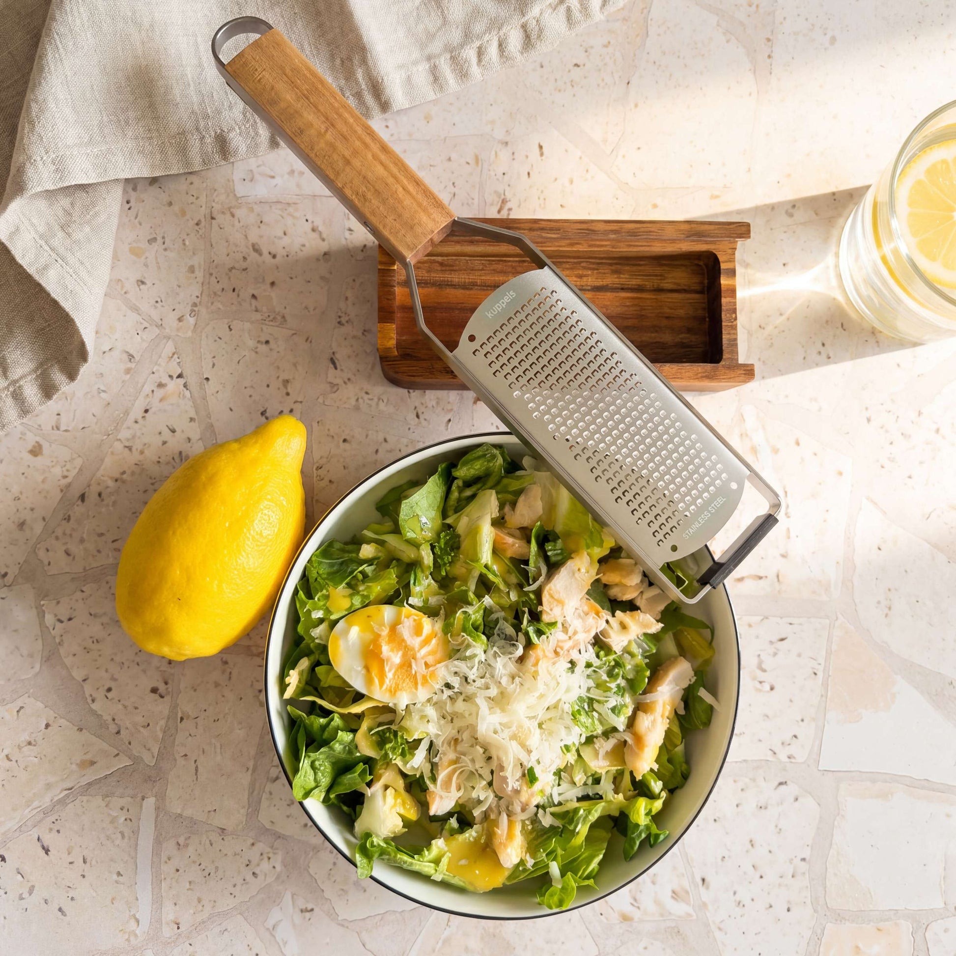 Eine Schüssel Salat mit geriebenem Käse, einem halbierten gekochten Ei und frischem Blattgemüse steht neben einer leuchtend gelben Zitrone, der Kuppels GRANO Handreibe „fein“ mit ihrem Holzbehälter und einem kleinen Akazienholz-Behälter. In der Ecke steht ein Glas Zitronenwasser auf einer hellen, strukturierten Oberfläche.