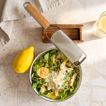 Eine Schüssel Salat mit geriebenem Käse, einem halbierten gekochten Ei und frischem Blattgemüse steht neben einer leuchtend gelben Zitrone, der Kuppels GRANO Handreibe „fein“ mit ihrem Holzbehälter und einem kleinen Akazienholz-Behälter. In der Ecke steht ein Glas Zitronenwasser auf einer hellen, strukturierten Oberfläche.