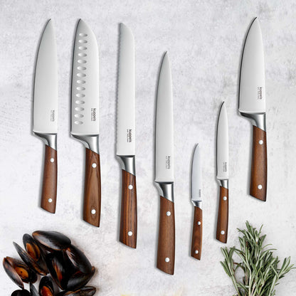 Sieben AVID Gemüsemesser von Kuppels, jeweils mit eleganten Griffen aus Eschenholz, sind in einer Reihe auf einer hellen Arbeitsplatte angeordnet; daneben liegen auf der linken Seite frischer Rosmarin und eine Schüssel mit Muscheln.